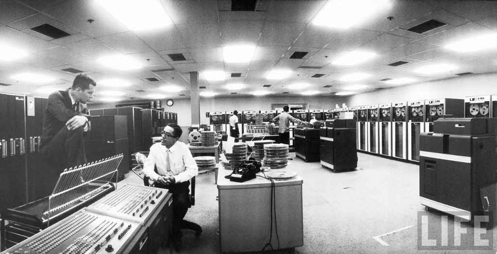  Robert Fairbanks and Robert Davis, both of IBM talking in computer room of North American Aviation. Magnetic tape drives and consoles and IBM computers are in the room.